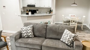 A modern living room with a grey sofa, two patterned pillows, a round coffee table, and a dining area with a white table and chairs.