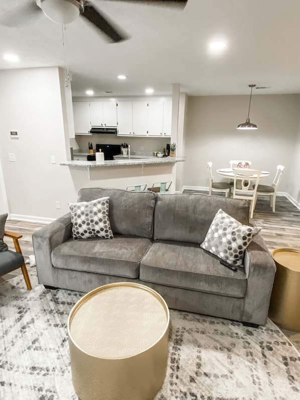 A modern living room with a grey sofa, two patterned pillows, a round coffee table, and a dining area with a white table and chairs.