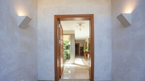 A hallway with a wooden door, light fixtures on the walls, and a tiled floor.