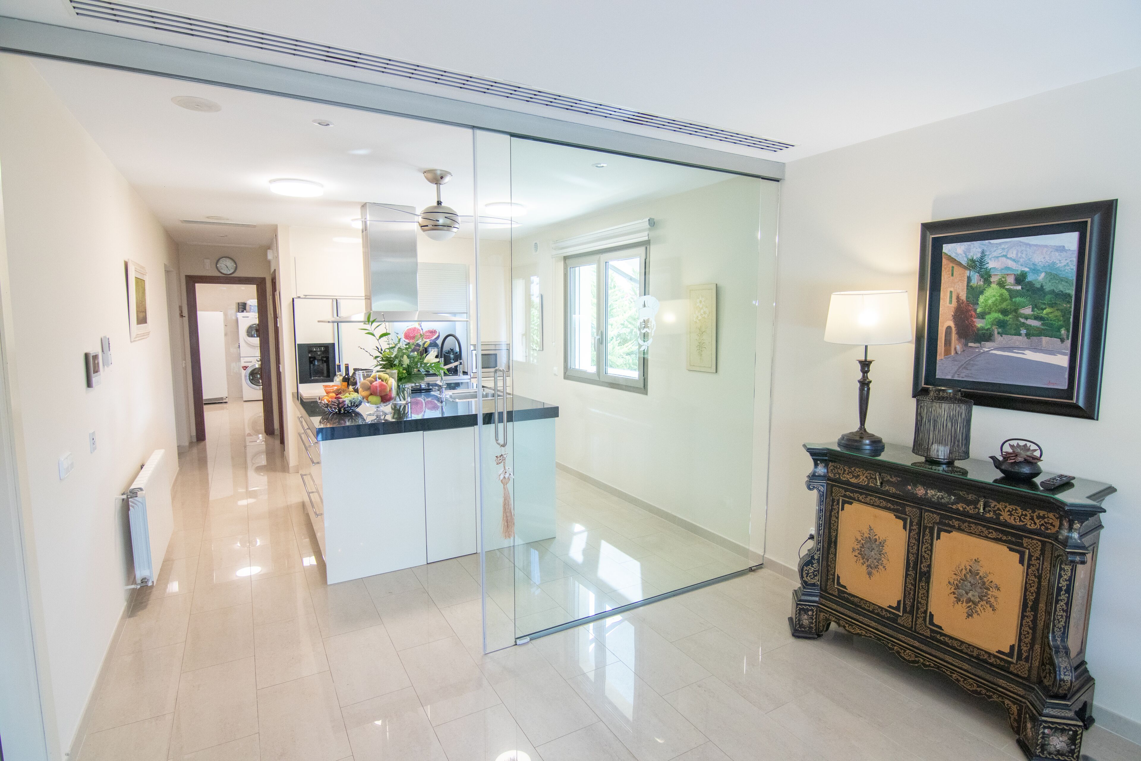 A modern kitchen with a glass partition, a central island, and a decorative cabinet with a lamp.