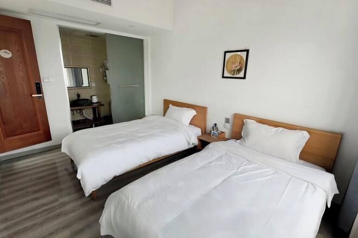 Hotellihuone, jossa on kaksi sänkyä, puinen ovi, pieni pöytä, jonka päällä on pullo, ja seinällä oleva kehystetty kuva.
