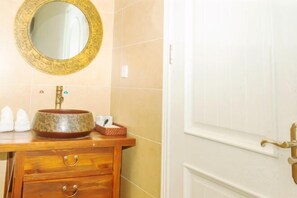 Un baño con tocador de madera, un espejo redondo con marco ornamentado y un lavabo con recipiente decorativo.