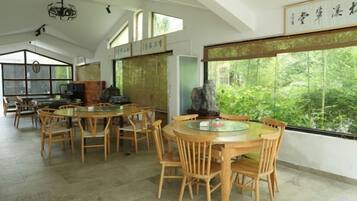 A dining area with round wooden tables and chairs, large windows with a view of greenery, and traditional decor.