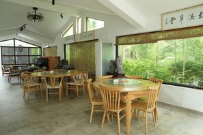 A dining area with round wooden tables and chairs, large windows with a view of greenery, and traditional decor.