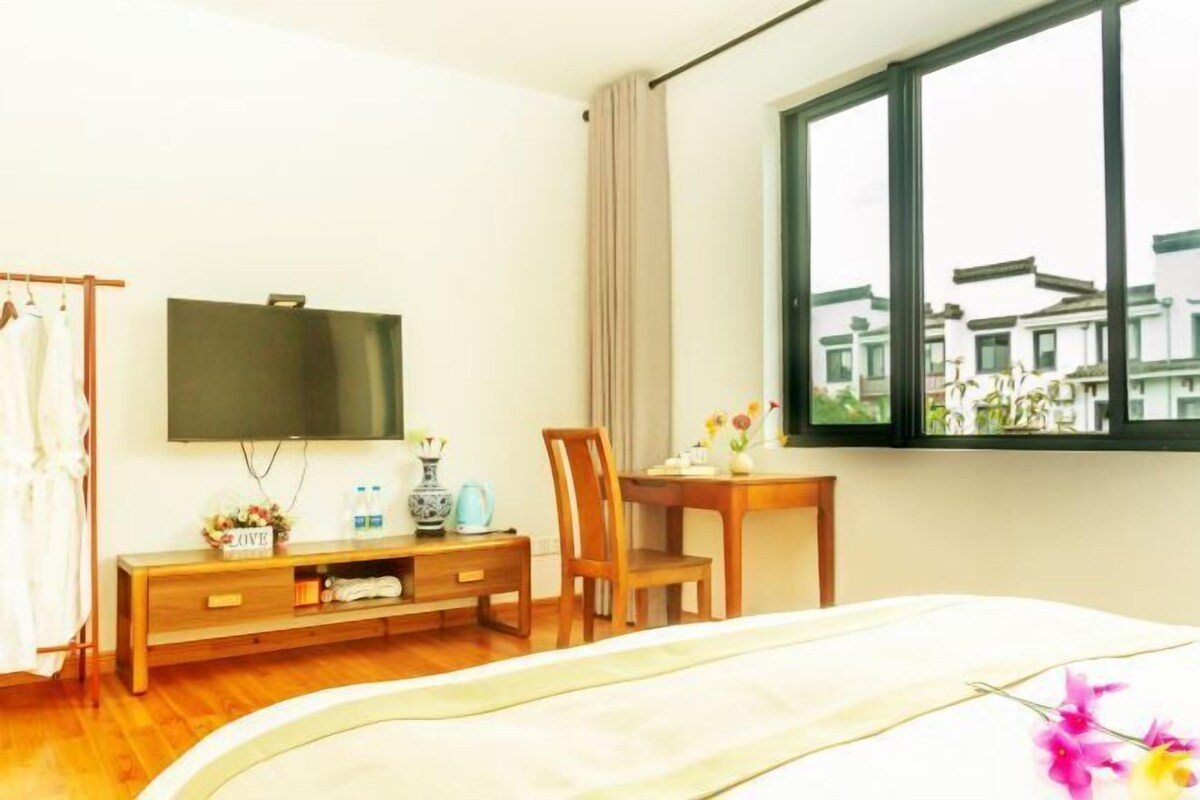 Habitación de hotel con cama, televisor montado en la pared, escritorio de madera, una silla y ventana con vistas a edificios.
