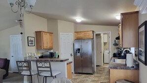 A kitchen with wooden cabinets, a stainless steel refrigerator, a dining area with a round table and bar stools, and a trash can. - Lake Roosevelt View Retreat Grand Coulee (Grand Coulee)