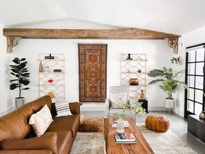 A living room with a brown leather sofa, a wooden coffee table, and a large patterned rug. There are two side tables with decorative items and a large window with a view. - Boho(asis) - Incredible Views, Dog Friendly, Quiet (La Quinta)