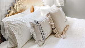 Un dormitorio con cama, cabecera de madera, ropa de cama blanca y una planta colgante.