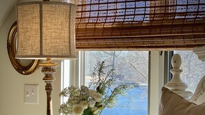 Un dormitorio con cama, mesita de noche con lámpara, jarra con flores, cuadro enmarcado y ventana con vista a árboles y una masa de agua.