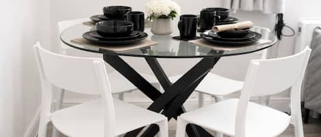 Una mesa redonda de vidrio con sillas blancas y negras, platos negros, tazas y un florero con flores blancas.