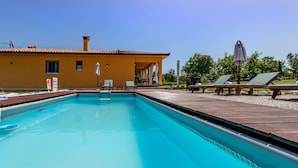 Une piscine avec des chaises longues et des parasols, un bâtiment jaune avec un toit en tuiles et un ciel bleu clair.
