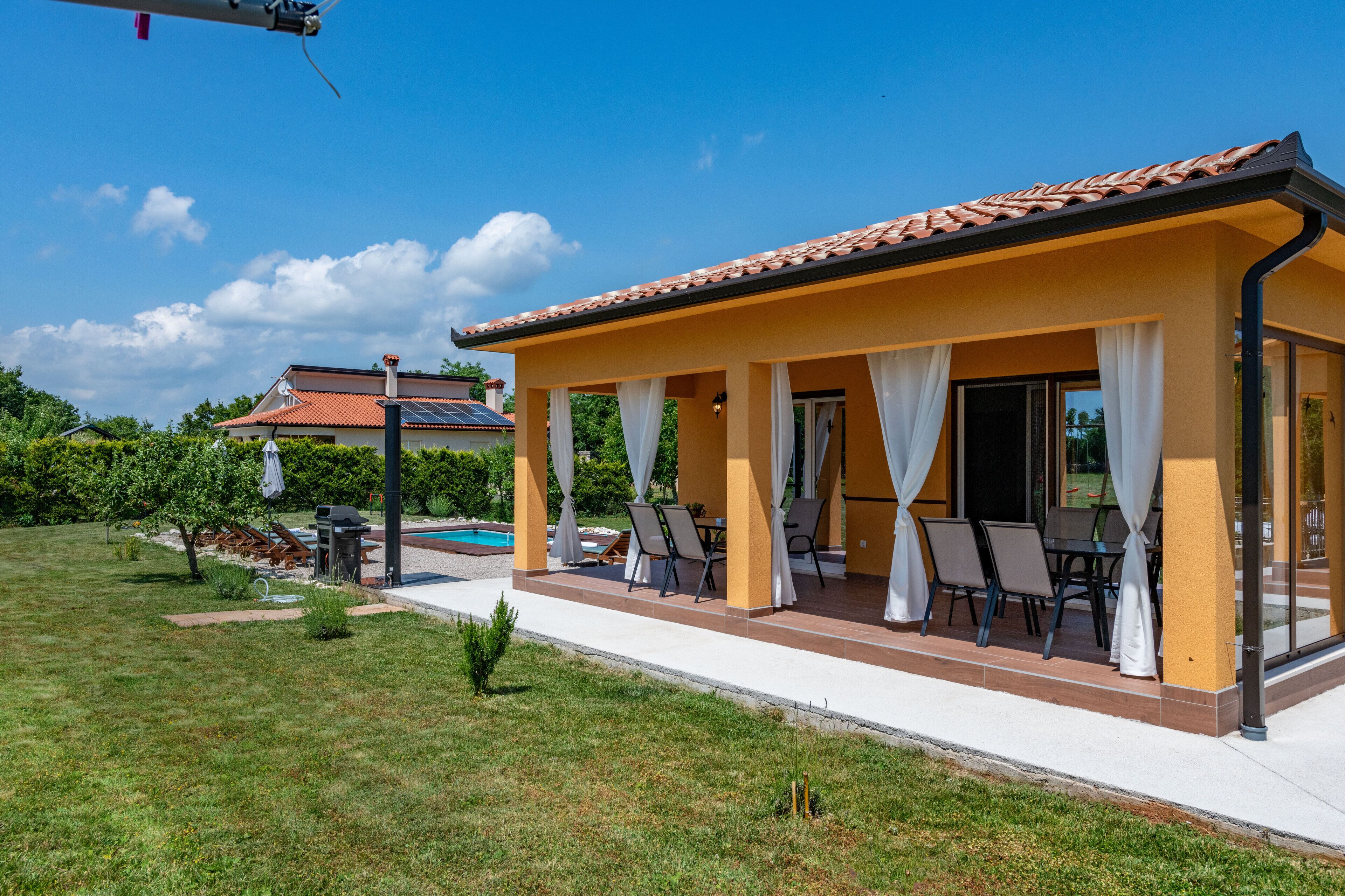 Une terrasse extérieure couverte, avec des chaises et une table, une piscine en arrière-plan et un ciel bleu dégagé.