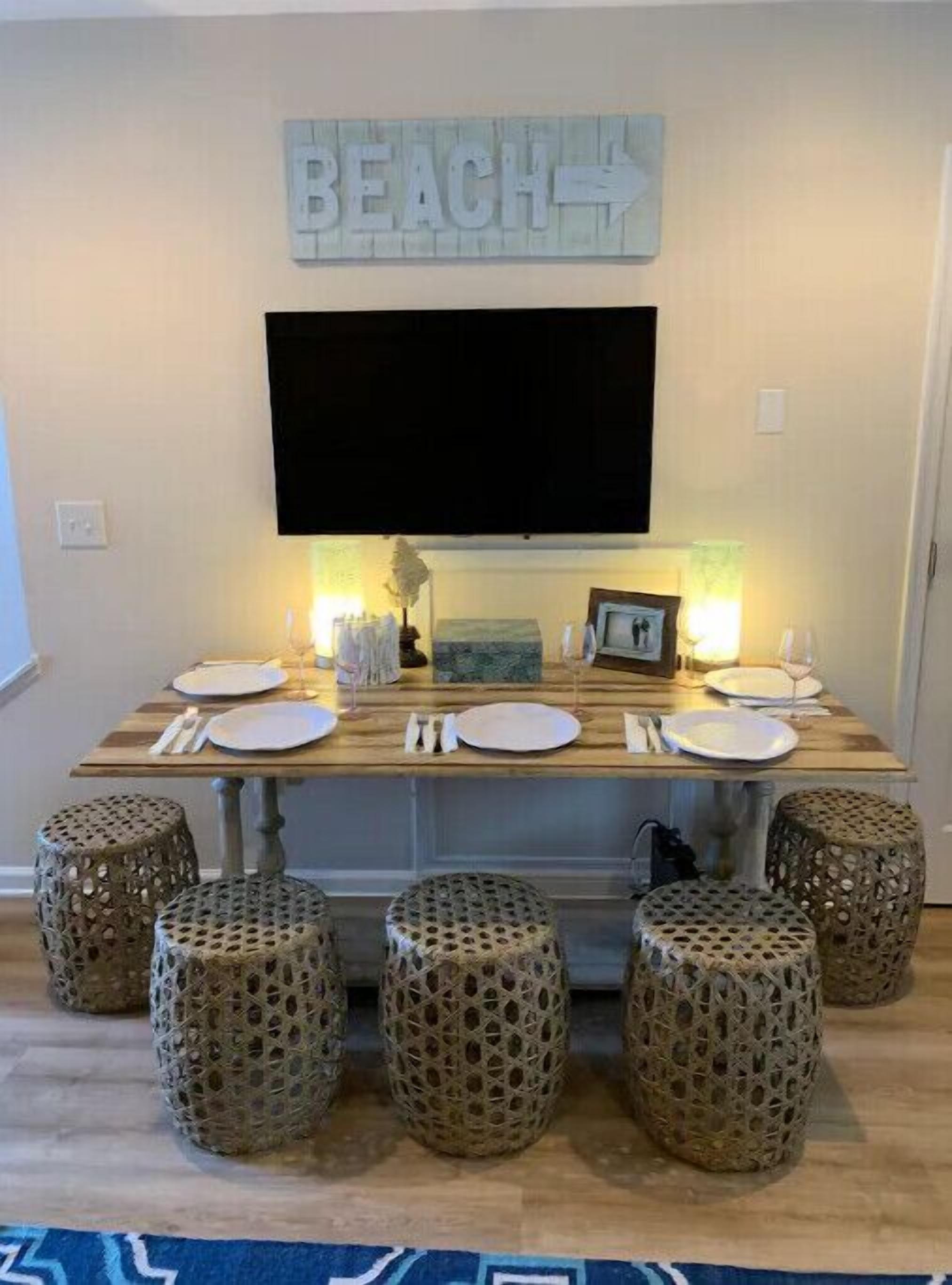 Una mesa para comer dispuesta con platos, cubiertos y vasos, rodeada de taburetes tejidos, con un letrero que dice 'BEACH' encima.