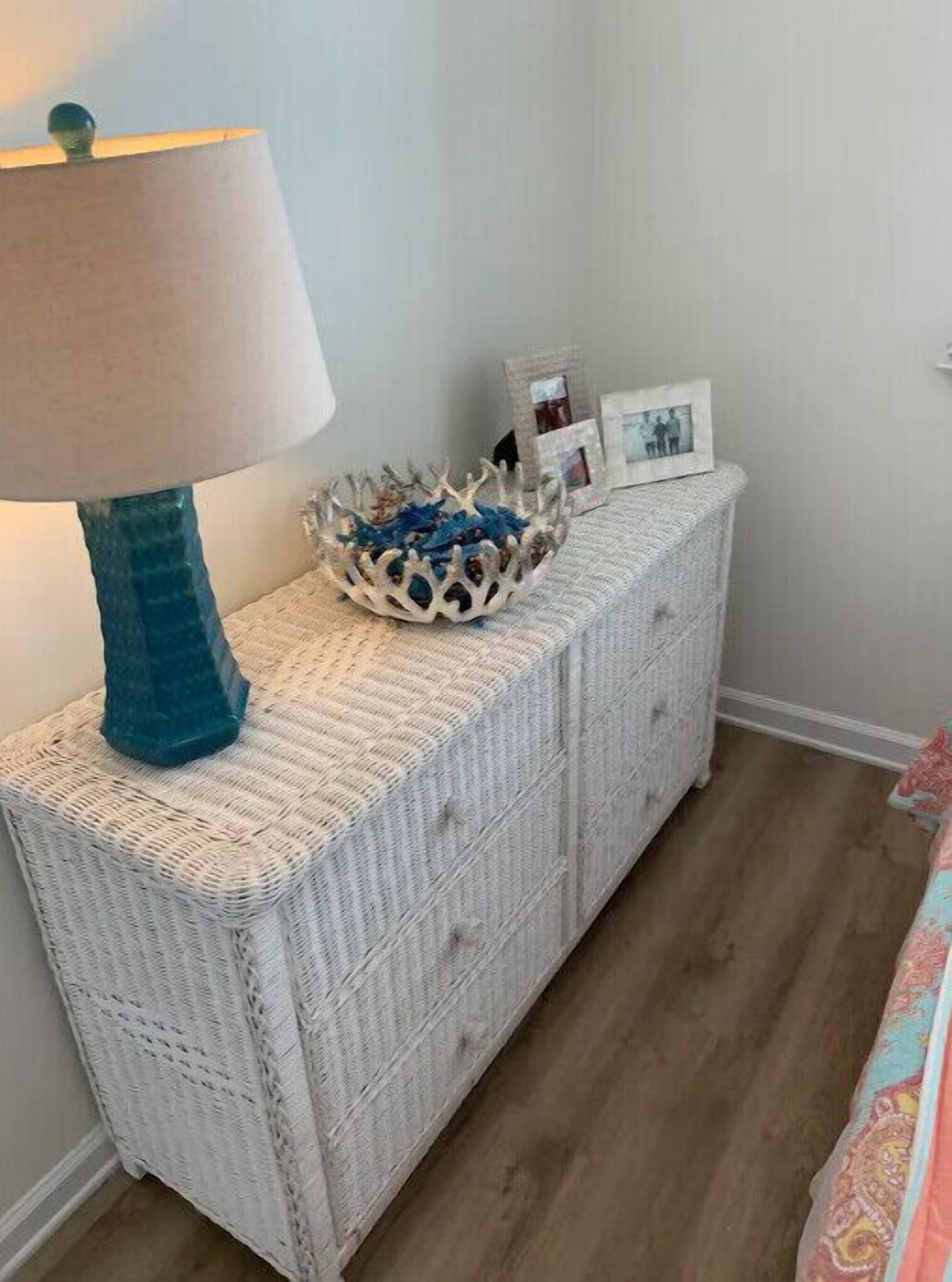 Una consola blanca de mimbre con una lámpara azul, un cuenco decorativo y fotos enmarcadas.