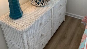 Een witte rieten console tafel met een blauwe lamp, een decoratieve schaal en ingelijste foto's.