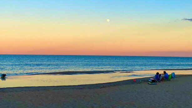 Een strandscène met een heldere lucht, een zichtbare maan en mensen die op het zand zitten.