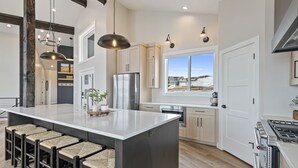 A spacious kitchen with a large island, wooden bar stools, stainless steel appliances, and a view of a snowy landscape through the window. - Highland Chateau at Longridge  (Sleeps 34) (Garden City)