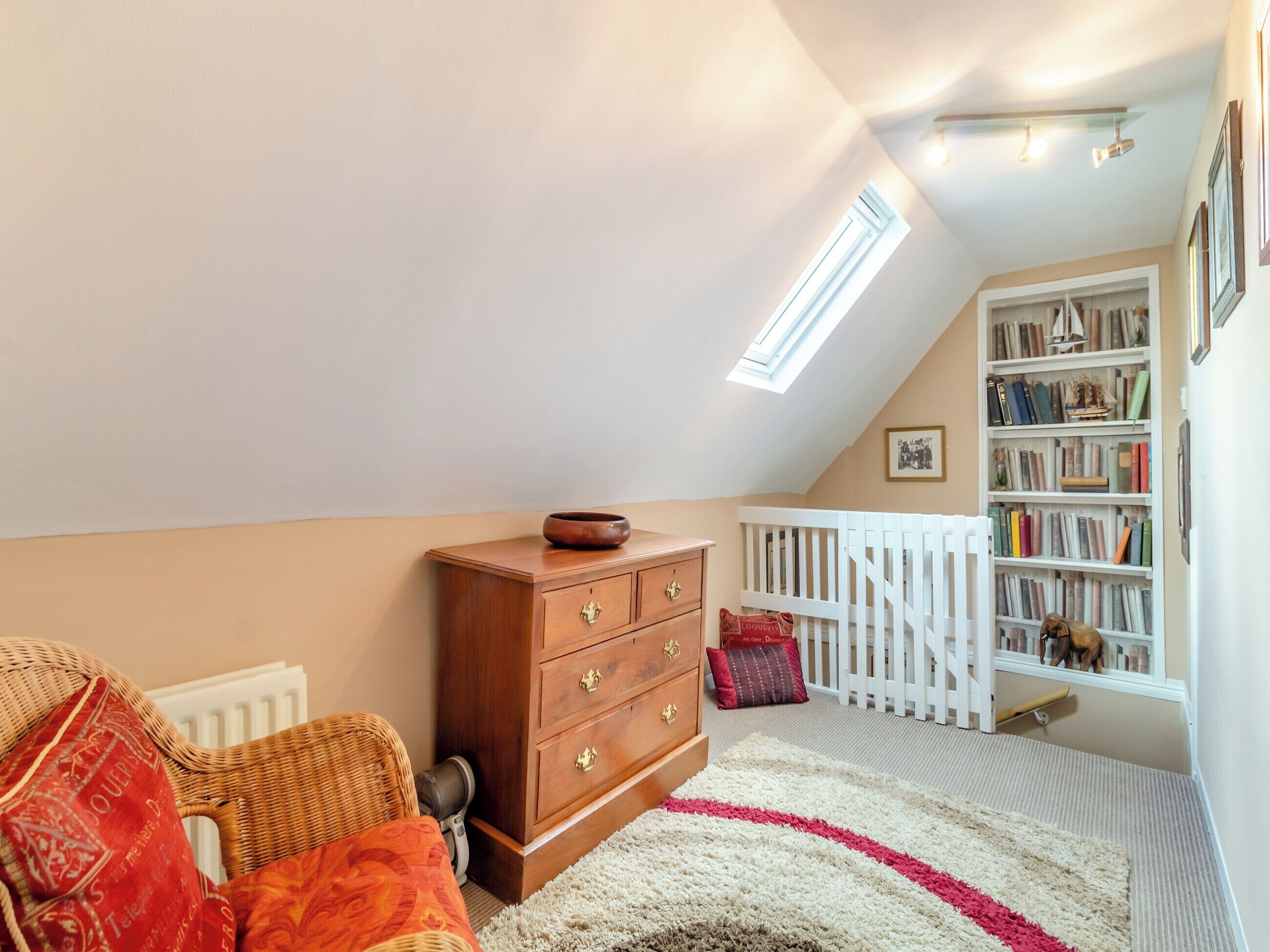 Ein gemütliches Schlafzimmer im Dachgeschoss mit schrägen Wänden, einer hölzernen Kommode, einem Korbstuhl, einem weißen Babybett und einem Bücherregal.