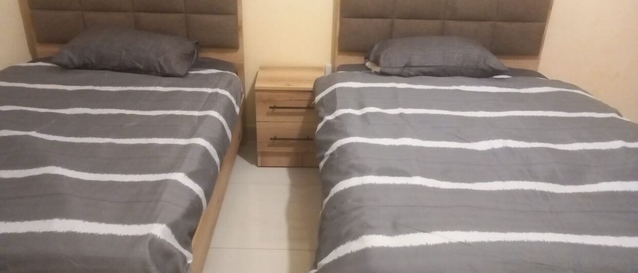 Deux lits jumeaux avec une literie à rayures, une table de chevet en bois avec des tiroirs et une tête de lit rembourrée.