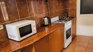 A kitchen with wooden cabinets, a white microwave, a kettle, and a stove. - RETRO PALACE (Tashkent)