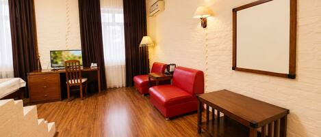 A hotel room with a wooden floor, a desk with a chair, a red sofa, a small table, a wall-mounted lamp, and a large mirror.