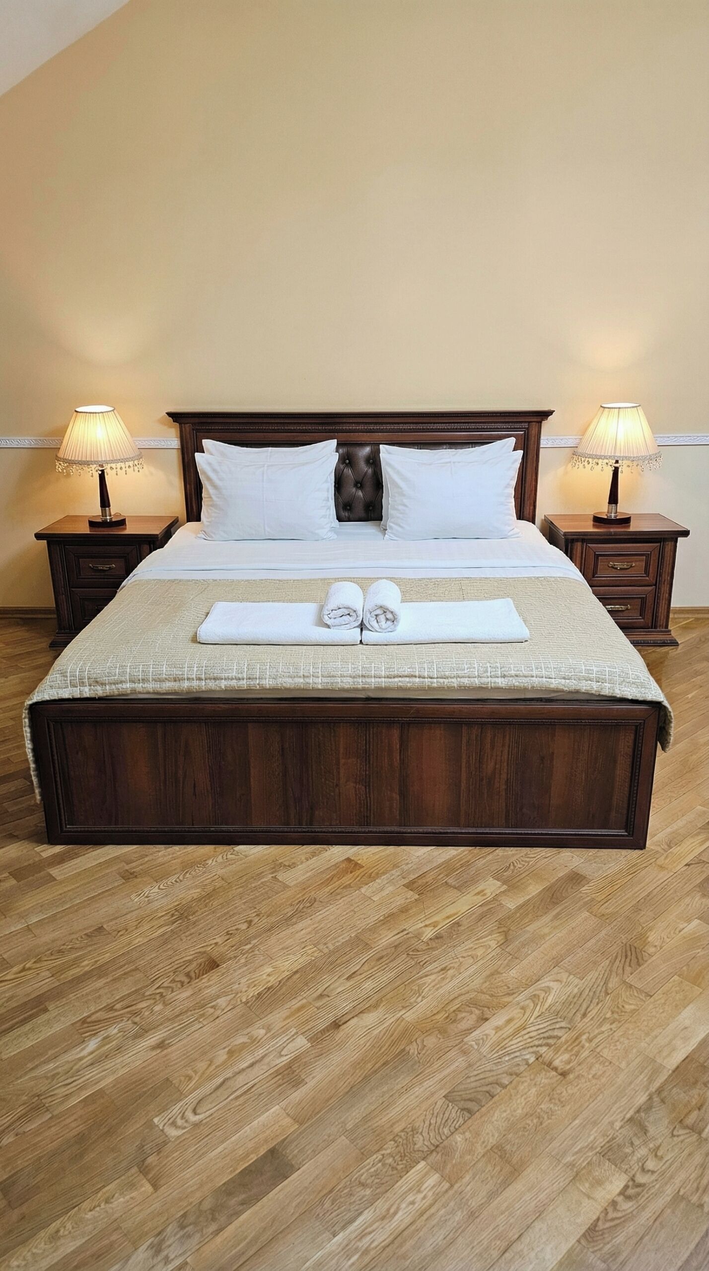 A neatly made bed with white linens and two rolled towels, flanked by two bedside tables with lamps, in a room with wooden flooring.