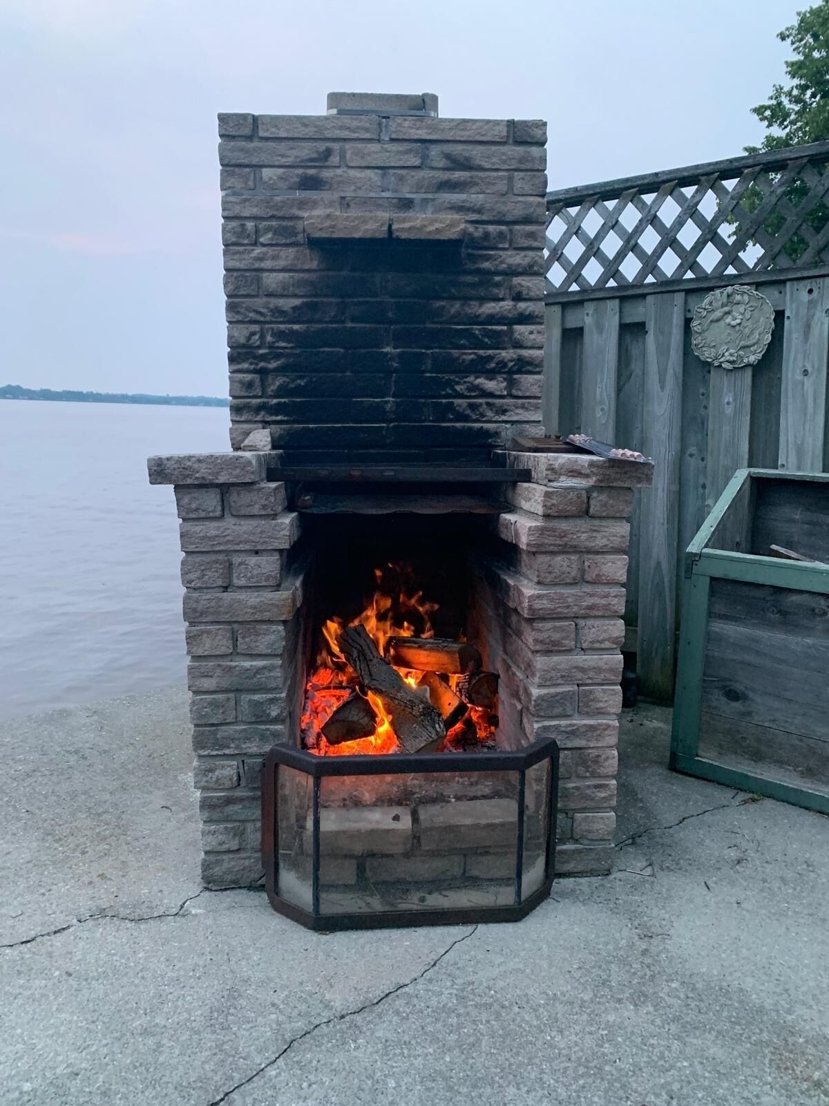 A stone-built outdoor fireplace with a fire burning inside, surrounded by a wooden fence and a view of a body of water.