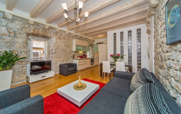 A modern living room with a stone wall, a flat-screen TV, grey sofas, a red rug, and a dining area with a glass partition.