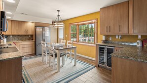 A modern kitchen with wooden cabinets, granite countertops, a dining table with chairs, and a large window with a view of greenery.