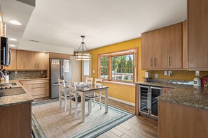 A modern kitchen with wooden cabinets, granite countertops, a dining table with chairs, and a large window with a view of greenery.