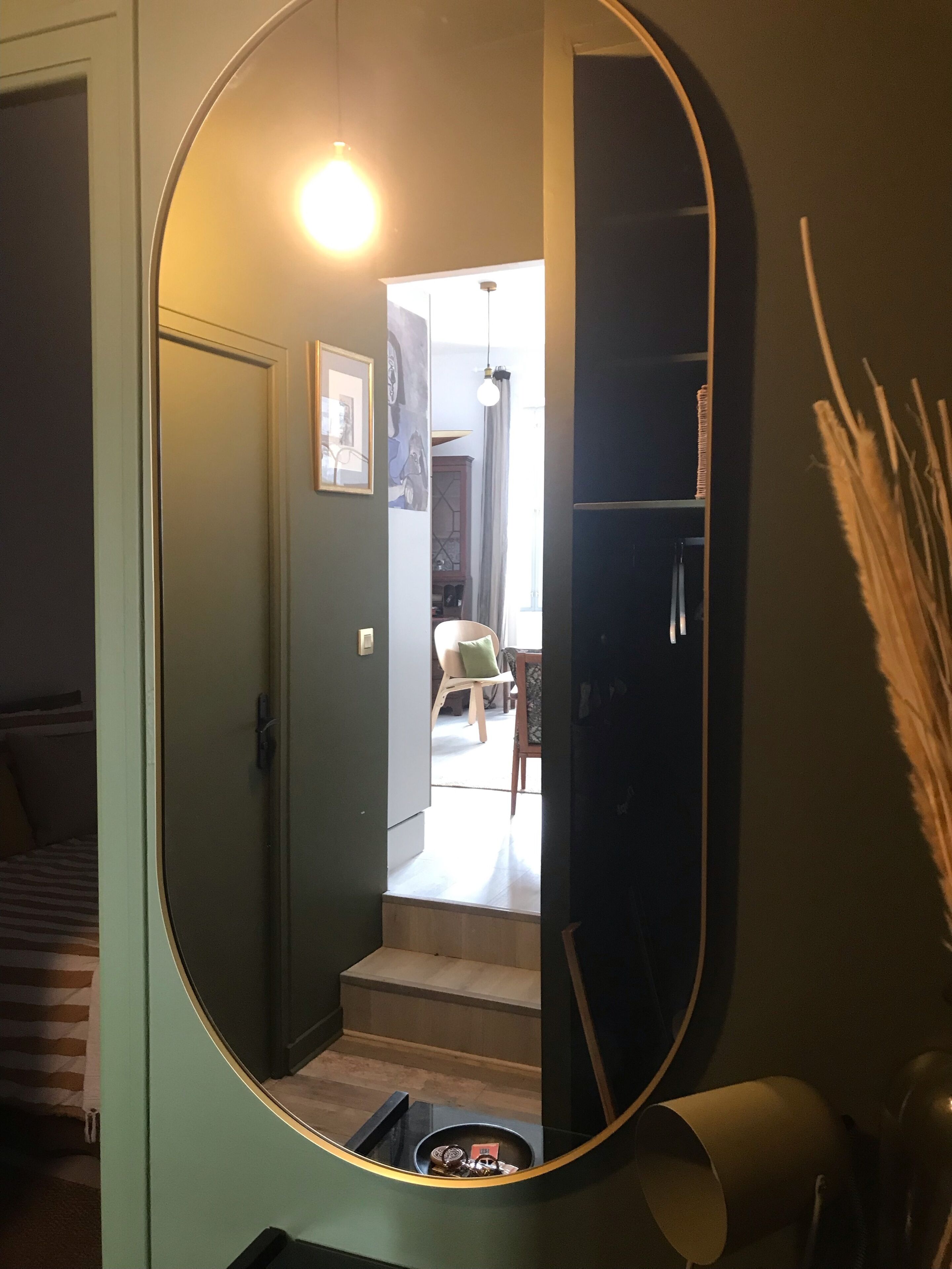 A hallway with a large mirror reflecting a room with a bed, a chair, and a staircase.