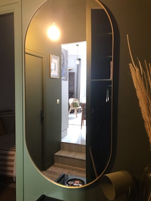 Un couloir avec un grand miroir qui reflète une pièce contenant un lit, une chaise et un escalier.
