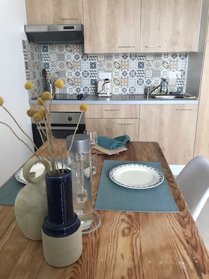 Une table de salle à manger en bois, avec une assiette, un vase contenant des fleurs séchées, une bouteille d’eau et un verre.