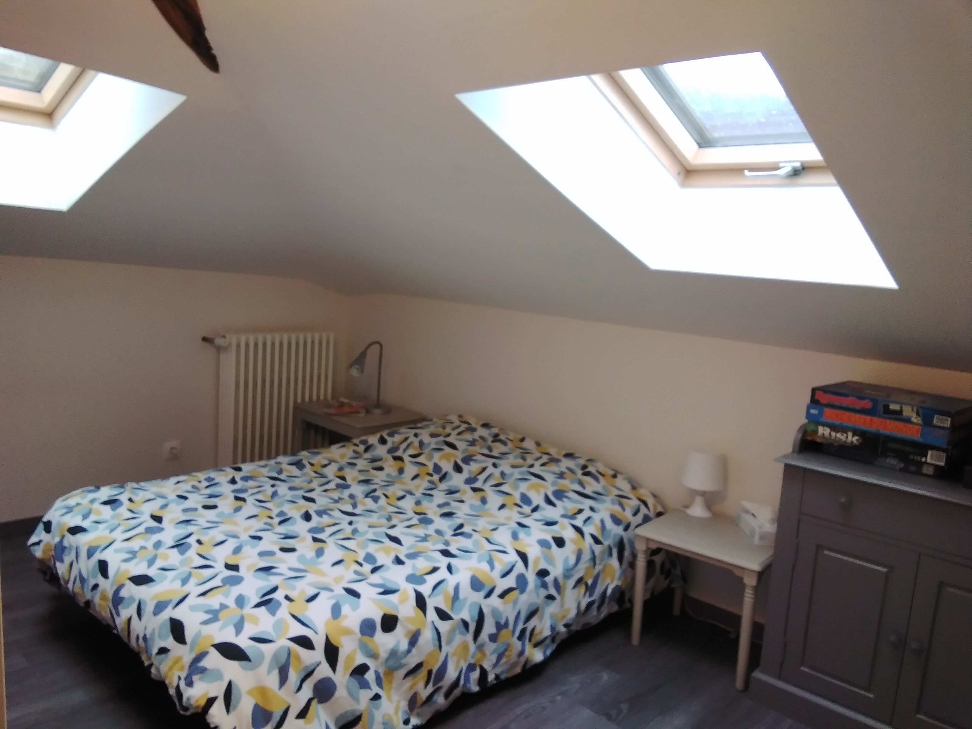 A small, well-lit room with a bed, a desk, and a cabinet. There are skylights on the ceiling, and a radiator is visible on the wall.