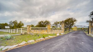 Una entrada con puerta de reja y un camino pavimentado que conduce a un edificio, rodeado de muros de piedra y una valla blanca.