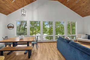 A spacious room with a high ceiling, large windows, a dining table with chairs, a blue sofa, and a hanging light fixture. - Lake Montcalm Waterfront Retreat - Beach, Hot Tub, Game Room (Six Lakes)