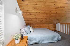 A bedroom with a wooden ceiling, a bed with a quilted cover, a bedside table with a plant, and a wall-mounted light fixture.