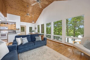 A spacious living area with a blue sofa, a kitchen with wooden cabinets, and large windows offering a view of trees.