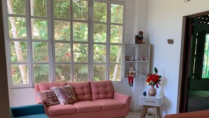 Una sala de estar con un sofá rosa, un sillón verde azulado y un sillón rojo. Se observa una estantería blanca con objetos decorativos y un jarrón de flores rojas.