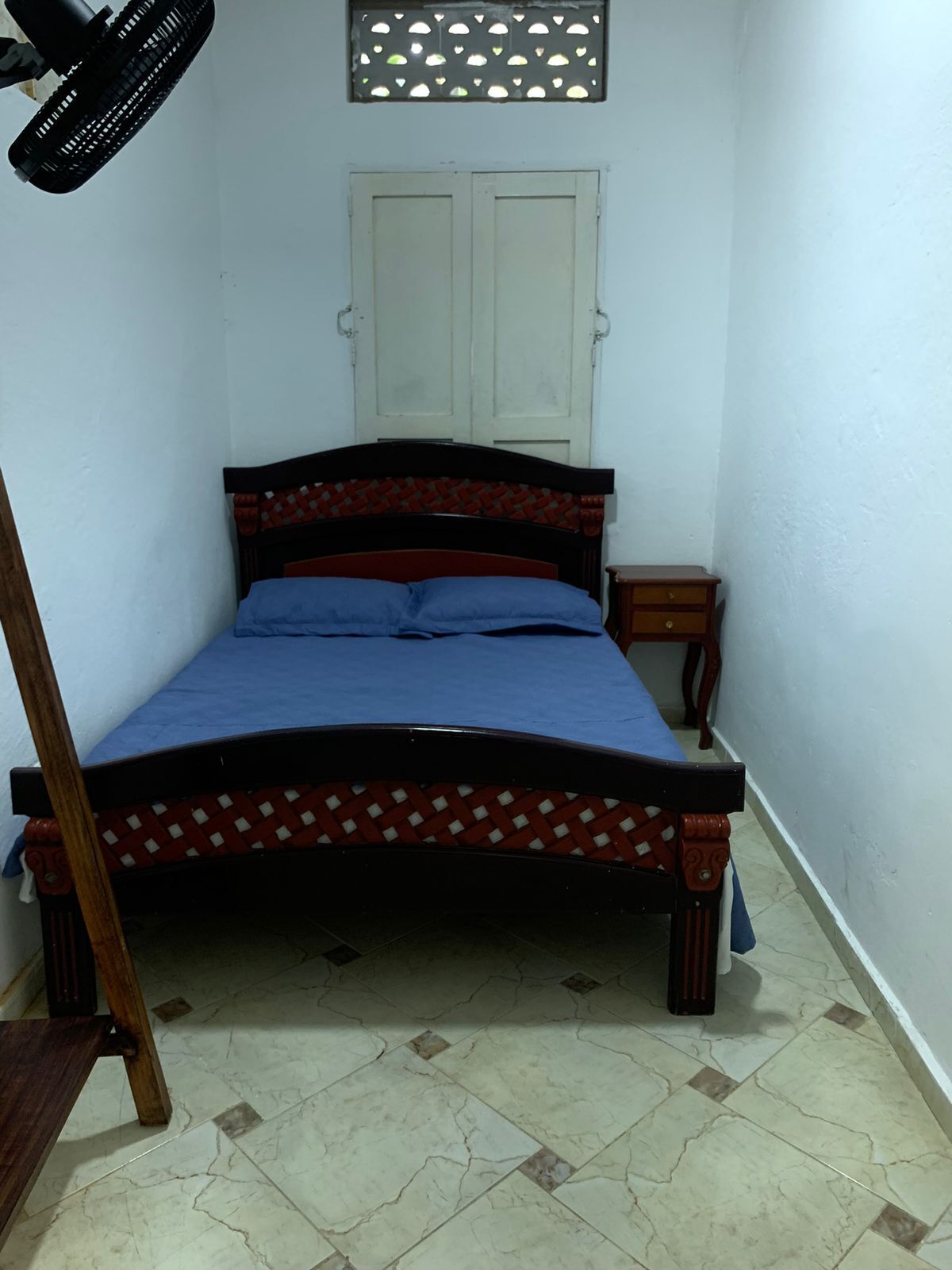 A small, narrow room with a bed, a wooden nightstand, a wall-mounted fan, and a window with decorative cutouts.
