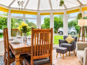 A conservatory with a wooden dining table set for four, surrounded by chairs, and a grey sofa with a cushion. - Synod Cottage | Private Hot Tub, Dog Friendly, Near New Quay (Ceredigion)