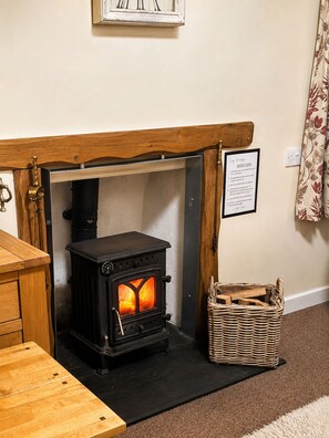 A wood-burning stove with a framed instruction sign above it, a wicker basket with firewood, and a wooden table. - Synod Cottage | Private Hot Tub, Dog Friendly, Near New Quay (Ceredigion)