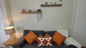 A living room with a grey sofa, two orange cushions, a patterned throw pillow, a small table with a lamp, and two wall-mounted shelves with decorative items and plants. - Pet-Friendly Country Escape HotTub & Log Burner (Ceredigion)