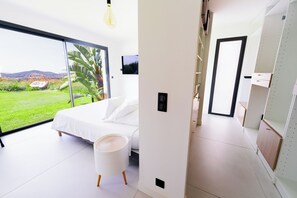 Quarto moderno com uma cama, uma pequena mesa de cabeceira branca e uma janela ampla com vista para uma paisagem verde.