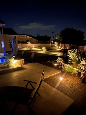 Uma área de varanda externa à noite, com piscina, assentos e plantas bem iluminadas.