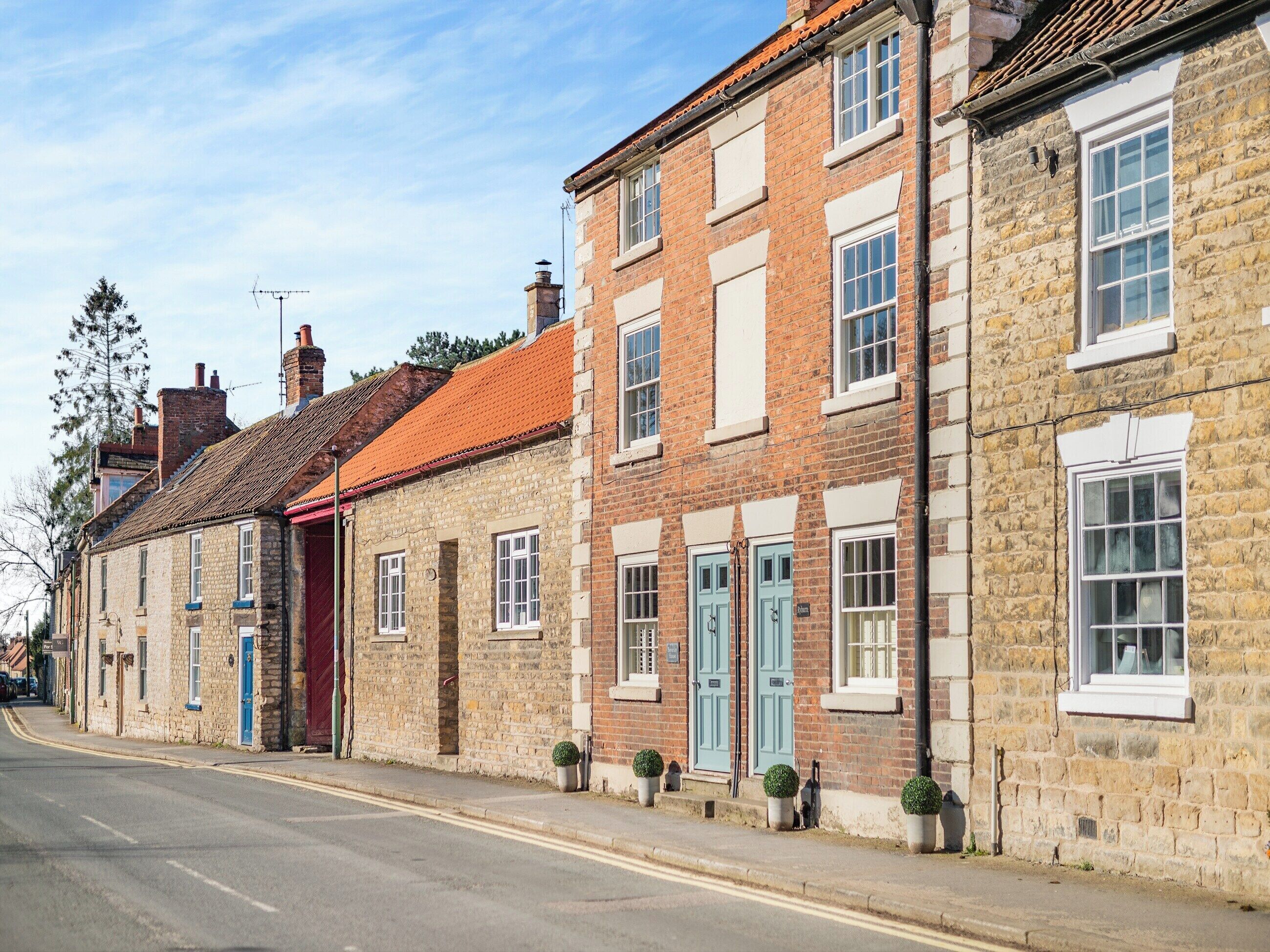 A row of traditional stone buildings with uniform window designs and doors, a clear road, and a tree on the left.