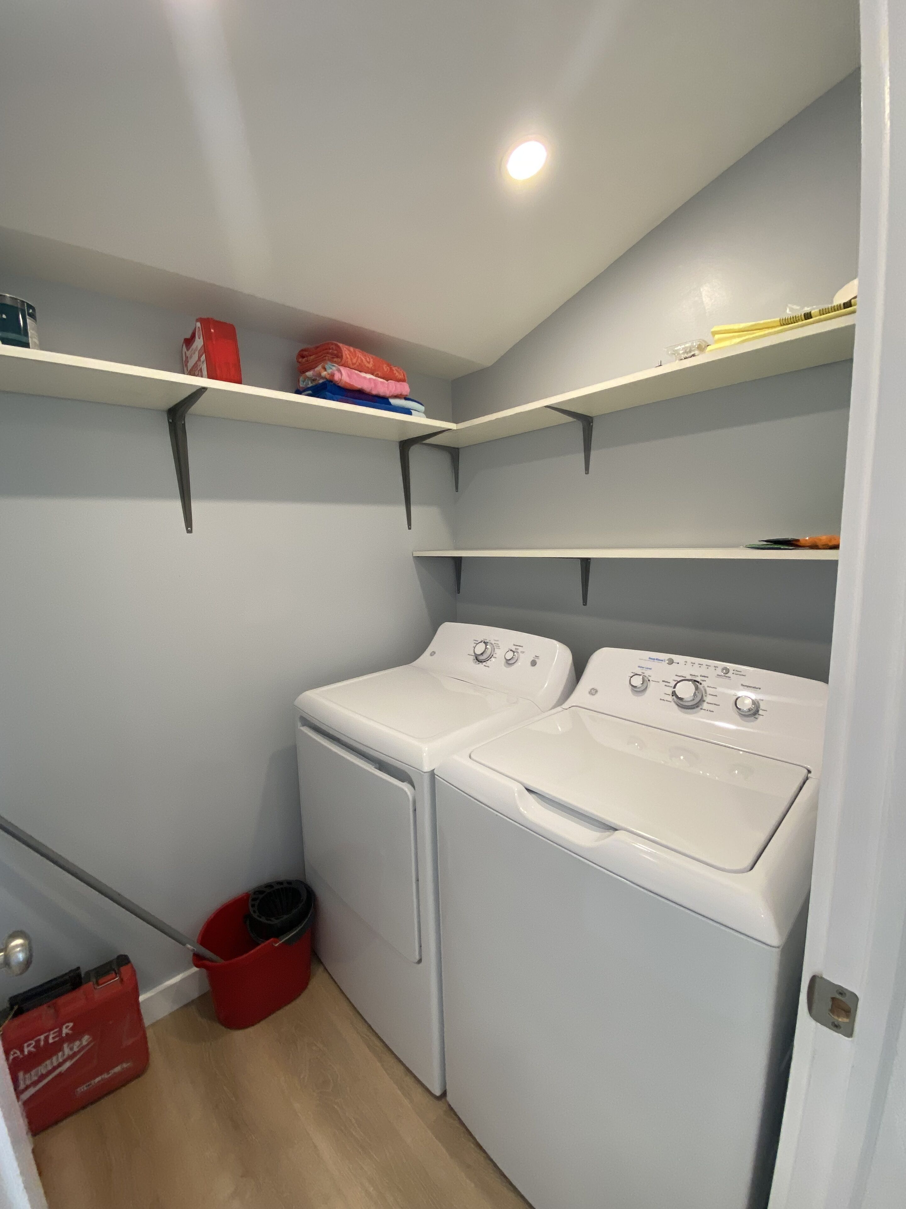 Une buanderie avec deux machines à laver blanches, un seau rouge et des étagères remplies de vêtements pliés et de produits de nettoyage.