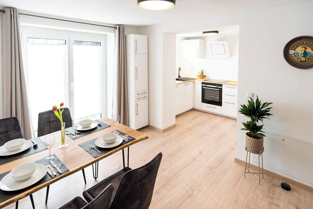 A modern kitchen with white cabinets, a dining area with a wooden table and chairs, and a potted plant.