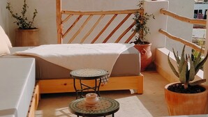 Een gezellig buitenverblijf met een houten bank, een klein rond tafeltje en een comfortabele zithoekbank. Er staan potplanten en er hangt een lamp.
