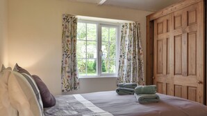 A neatly made bed with floral-patterned bedding, a window with curtains, and a wooden door. - 1 bedroom accommodation in Lostwithiel (Lostwithiel)